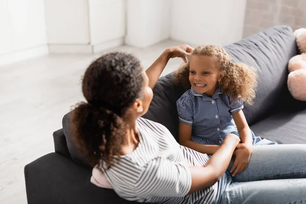 Vue grand angle de la mère touchant les cheveux de fille afro-américaine excitée tout en étant assis sur le canapé — Photo de stock