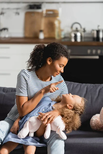Young african american mother in striped t-shirt having fun with daughter while sitting on sofa in kitchen — Stock Photo