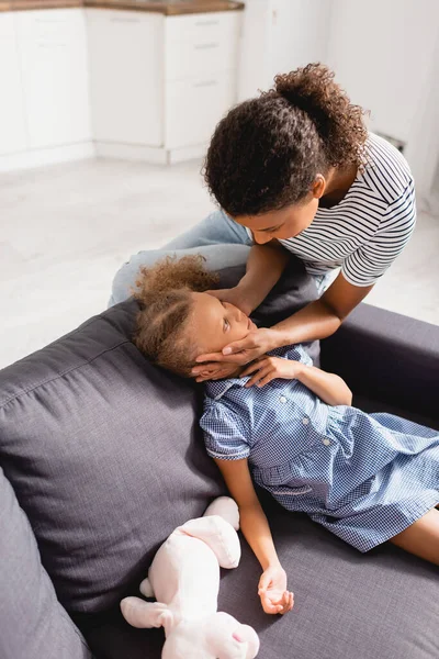 High angle view of african american woman touching face of daughter sitting on couch in dress near toy bunny — Stock Photo