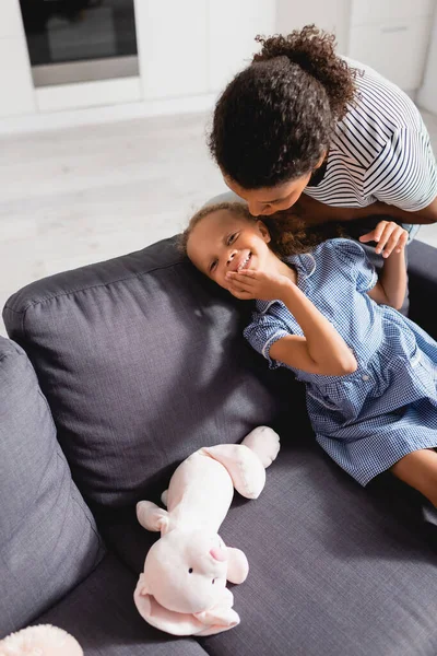 Africano americano mujer susurrando en el oído de excitado hija tocando cara mientras sentado en sofá cerca de juguete conejito - foto de stock