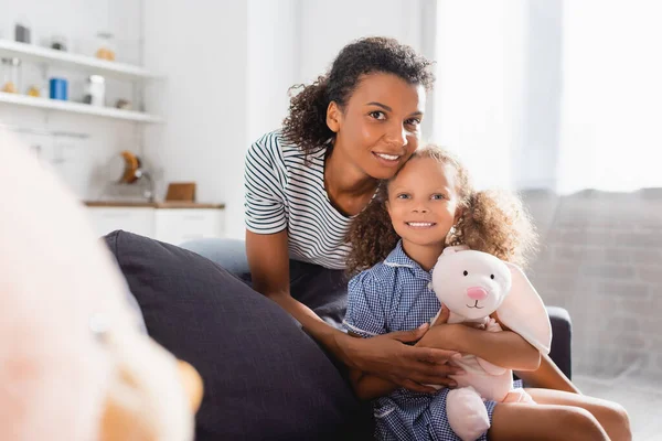 Selective focus of african american woman in striped t-shirt, and daughter with toy bunny looking at camera — Stock Photo