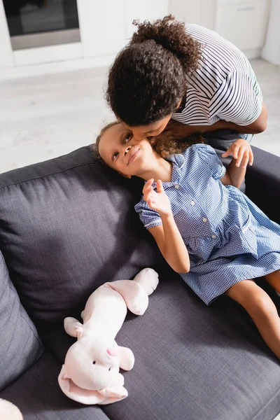 Vista ad alto angolo di donna afro-americana baciare figlia seduta sul divano in abito vicino coniglietto giocattolo — Foto stock