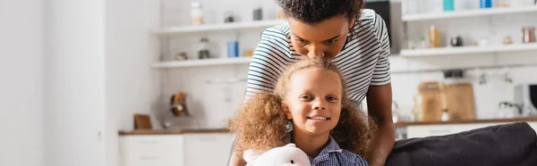 Panoramic concept of african american woman kissing head of daughter looking at camera in kitchen — Stock Photo