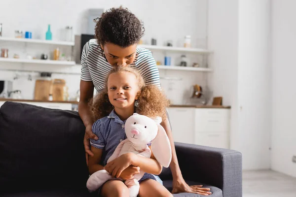 Junge afrikanisch-amerikanische Mutter küsst Kopf eines aufgeregten Kindes mit Spielzeughase und blickt in die Kamera — Stockfoto