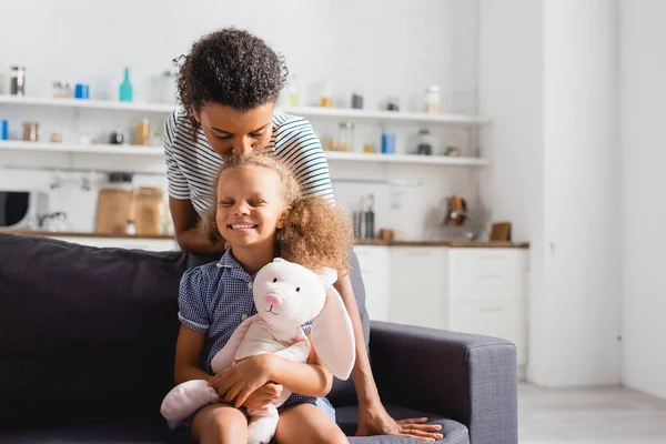 Junge afrikanisch-amerikanische Frau küsst Kopf der aufgeregten Tochter mit Spielzeughasen — Stockfoto