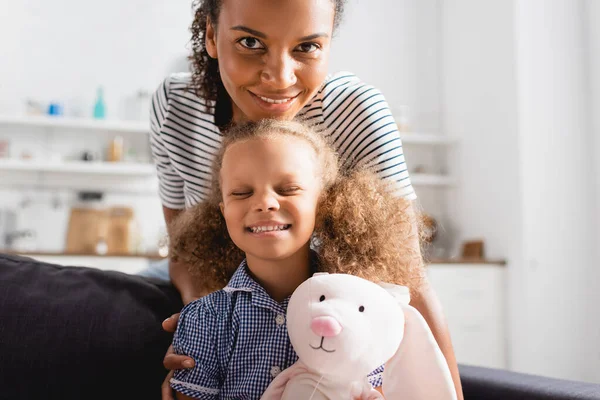 Junge afrikanisch-amerikanische Mutter blickt in die Kamera nahe aufgeregter Tochter mit geschlossenen Augen, die Spielzeughasen hält — Stockfoto