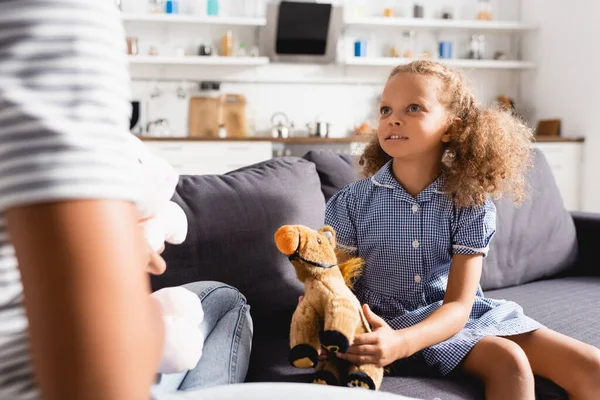 Cropped view of babysitter near african american girl in dress holding toy horse while sitting on sofa, selective focus — Stock Photo