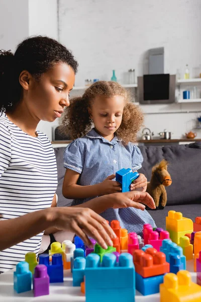 Enfoque selectivo de niñera afroamericana en camiseta a rayas y niña en vestido jugando con bloques de construcción de colores en casa - foto de stock