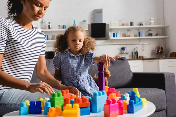 Afro-américaine fille en robe et baby-sitter en t-shirt rayé jouer avec des blocs de construction colorés dans la cuisine — Photo de stock