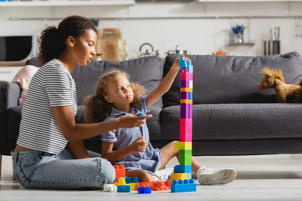 Africano americano ragazza costruzione torre da blocchi colorati mentre gioca sul pavimento vicino a giovane babysitter — Foto stock