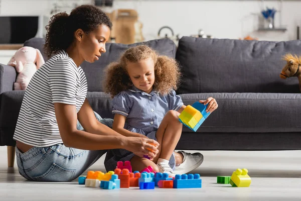 Chica afroamericana y niñera jugando con bloques de construcción de colores en el piso cerca del sofá - foto de stock