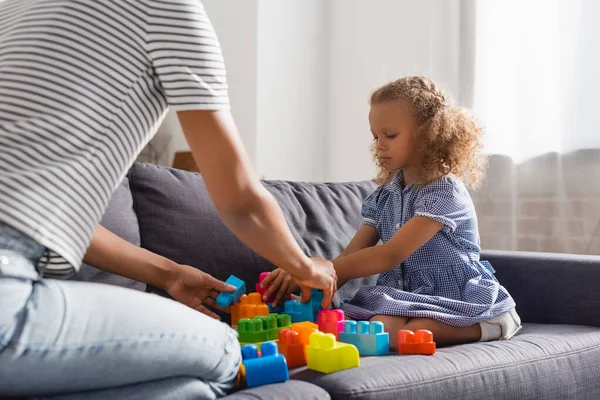 Vue partielle de baby-sitter et afro-américaine fille jouer avec des blocs de construction colorés sur le canapé — Photo de stock