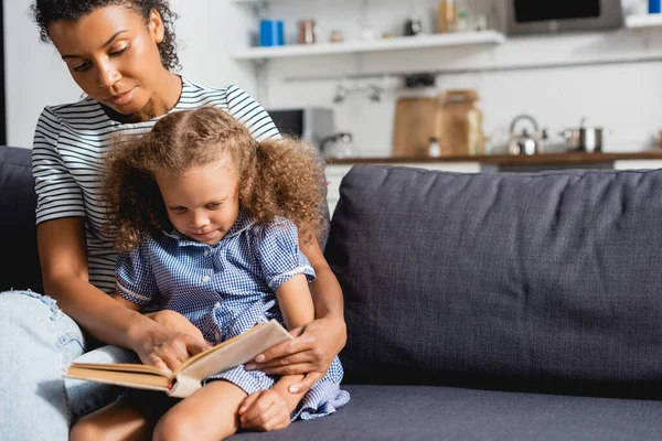 Joven afroamericano niñera y concentrado niño sentado en sofá y lectura libro juntos - foto de stock