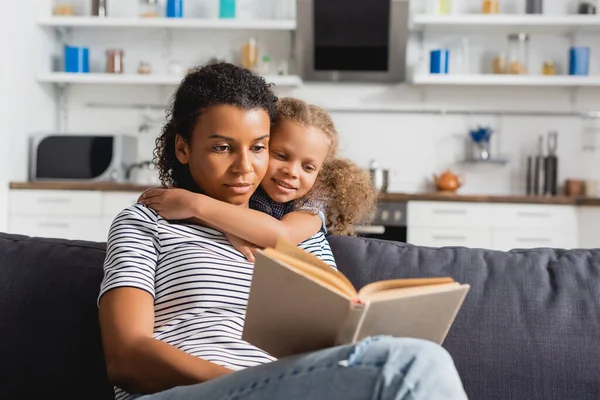 Konzentrierte afrikanisch-amerikanische Babysitterin im gestreiften T-Shirt liest Buch, während Mädchen ihren Hals umarmt — Stockfoto