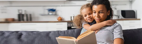 Imagen horizontal de niña afroamericana abrazando niñera sentada en el sofá y leyendo libro en la cocina - foto de stock