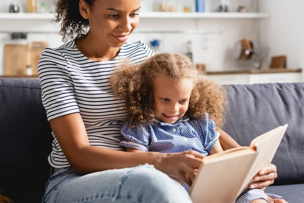 Foco seletivo de afro-americano babá e menina sentados no sofá e ler livro juntos — Fotografia de Stock