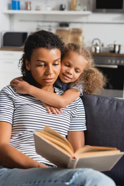 Enfoque selectivo de la niña afroamericana abrazando niñera en rayas libro de lectura de camisetas en el sofá - foto de stock