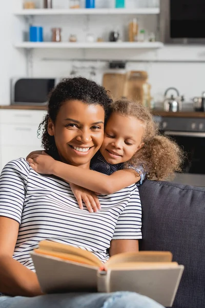 Selektiver Fokus der afrikanisch-amerikanischen Kindermädchen, die Buch in der Hand halten und in die Kamera schauen, während das Kind ihren Hals umarmt — Stockfoto