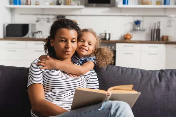 Afrikanisches amerikanisches Mädchen blickt in die Kamera und umarmt Kindermädchen, das auf der Couch sitzt und Buch liest — Stockfoto