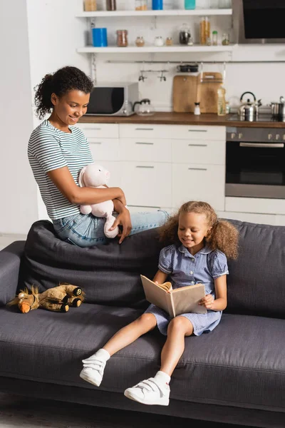 Молода афроамериканська няня тримає іграшкового кролика і дивиться на дівчину, читаючи книгу на дивані на кухні — стокове фото