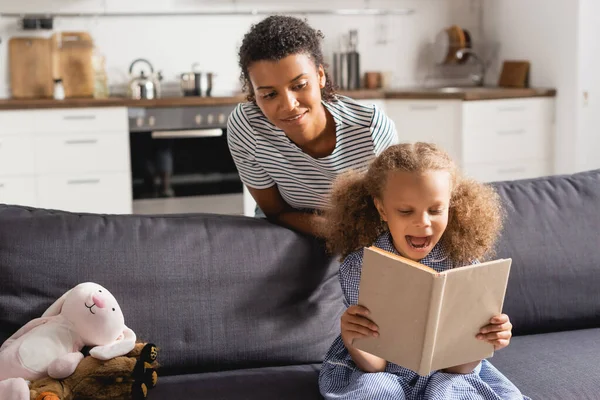 Jeune baby-sitter afro-américaine derrière enfant excité reding livre sur le canapé dans la cuisine — Photo de stock