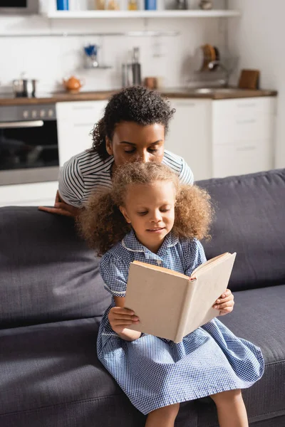 Afro americano donna con gli occhi chiusi baciare testa di figlia lettura libro su divano — Foto stock