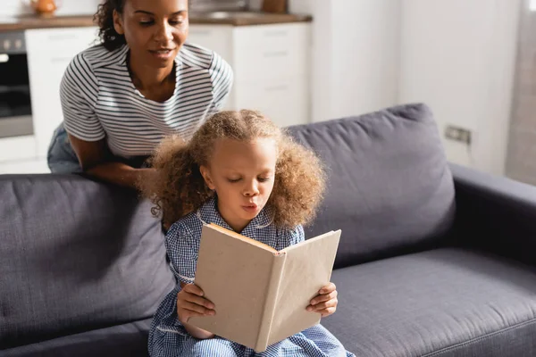 Niñera afroamericana en camiseta a rayas mirando a una chica concentrada sentada en un sofá y leyendo un libro - foto de stock