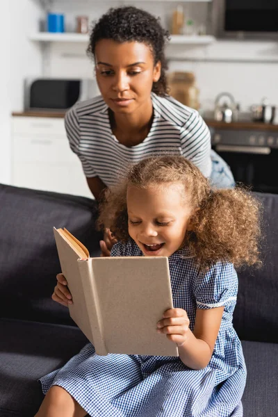 Jovem afro-americano babá em listrado t-shirt atrás animado menina leitura livro no sofá — Fotografia de Stock