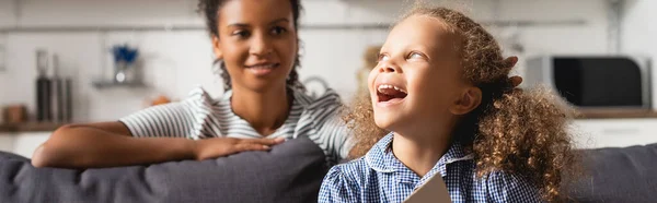 Enfoque selectivo de la niñera afroamericana joven detrás de la chica emocionada riendo mientras mira hacia otro lado, imagen horizontal - foto de stock