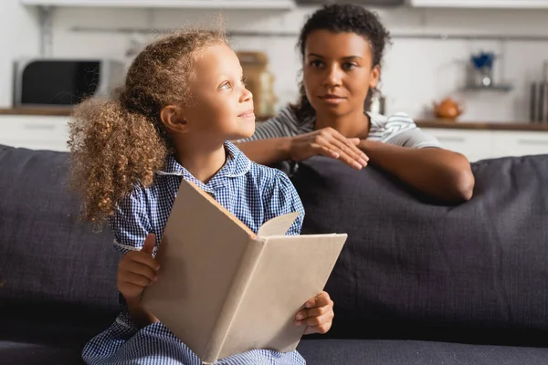 Enfoque selectivo de niñera afroamericana detrás de niño soñador mirando hacia otro lado mientras sostiene libro abierto - foto de stock