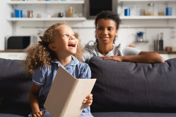 Foyer sélectif de babysitter afro-américain derrière fille excitée tenant livre ouvert tout en étant assis sur le canapé — Photo de stock