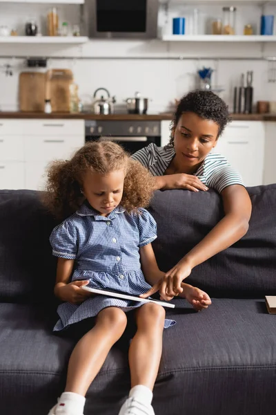 Afrikanische amerikanische Kindermädchen berühren digitales Tablet in den Händen von Mädchen, die auf dem Sofa sitzen — Stockfoto