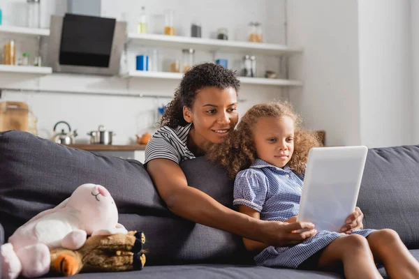 Niñera afroamericana joven sosteniendo tableta digital delante de un niño sentado en un sofá en la cocina - foto de stock