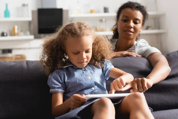 Foyer sélectif de nounou afro-américaine pointant du doigt la tablette numérique dans les mains de l'enfant assis sur le canapé — Photo de stock