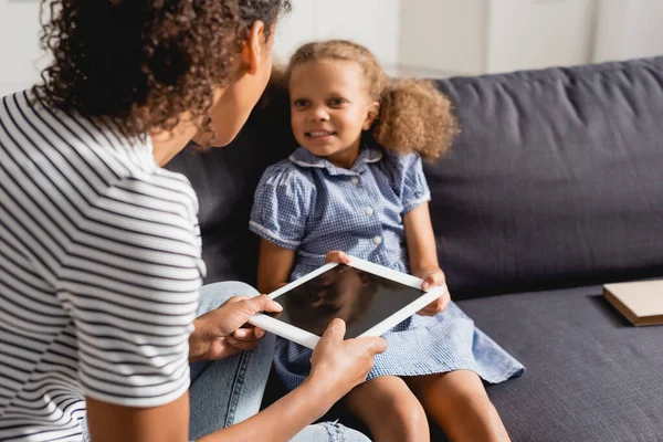 Foco seletivo de babá dando tablet digital com tela em branco para a menina afro-americana sentada no sofá — Fotografia de Stock