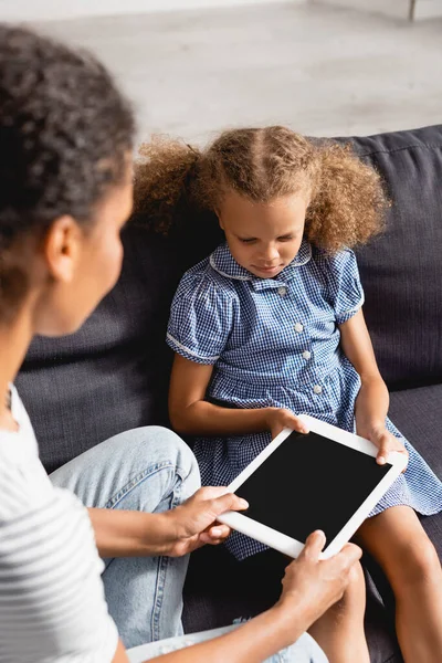 Enfoque selectivo de niñera dando tableta digital con pantalla en blanco a la chica afroamericana sentada en el sofá - foto de stock