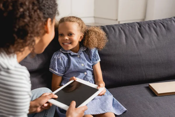 Enfoque selectivo de la niñera sosteniendo tableta digital con pantalla en blanco cerca de chica afroamericana sentada en el sofá - foto de stock