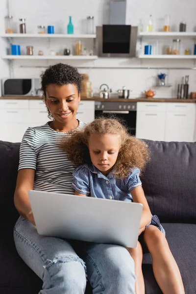 Joven afroamericano freelancer trabajando en portátil mientras está sentado con su hija en el sofá en la cocina - foto de stock