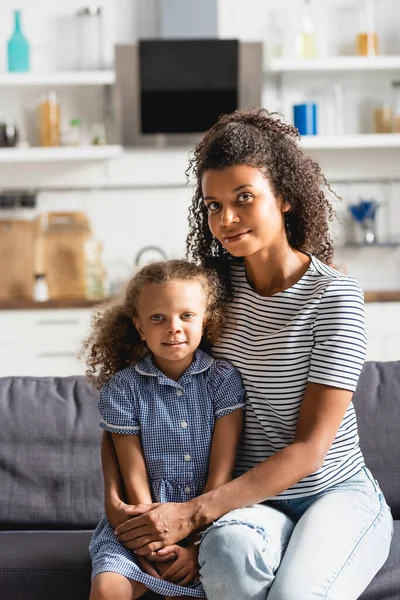Joven afroamericana americana mujer en rayas camiseta abrazando hija en vestido mientras mira la cámara - foto de stock