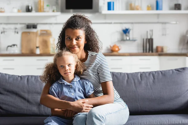 Молода афроамериканка обіймає дочку і дивиться на камеру, сидячи на дивані на кухні — стокове фото