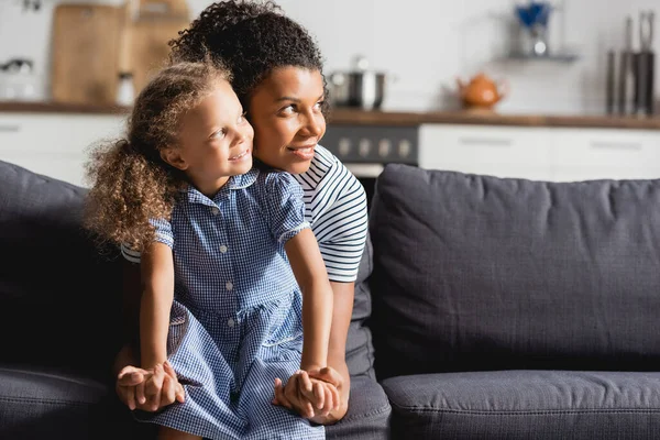 Mujer afroamericana y su hija tomados de la mano mientras están sentados en el sofá y mirando hacia otro lado - foto de stock
