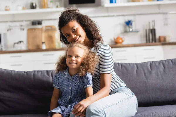 Africano americano madre y hija sentado en sofá en cocina y mirando cámara - foto de stock