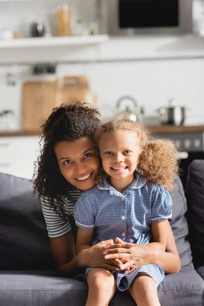 Jeune mère afro-américaine embrassant sa fille assise sur le canapé et regardant la caméra — Photo de stock