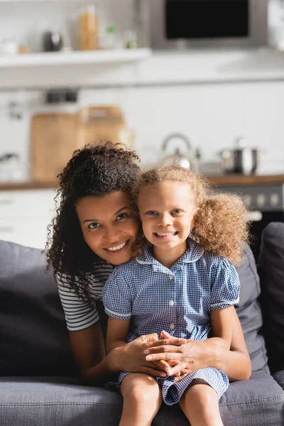 Giovane madre afroamericana che abbraccia la figlia seduta sul divano e guarda la macchina fotografica — Foto stock