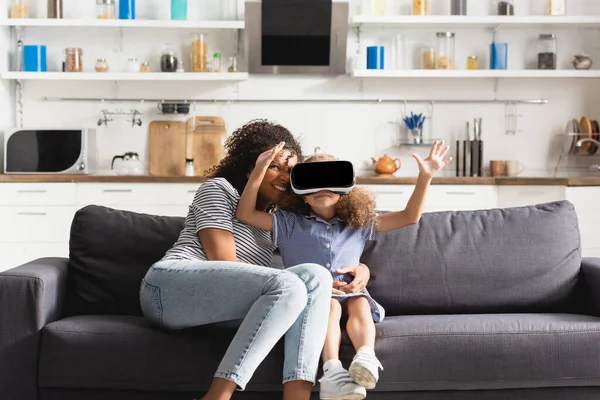 Junge afrikanisch-amerikanische Kindermädchen umarmt Kind in vr Headset gestikulierend auf Sofa in der Küche — Stockfoto