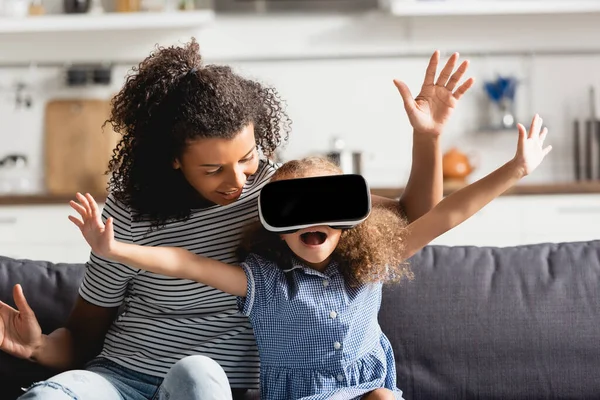 Young african american nanny and excited girl in vr headset gesturing with outstretched hands on couch — Stock Photo