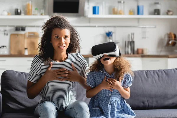 Schockierter afrikanisch-amerikanischer Babysitter und aufgeregtes Mädchen im Kopfhörer, das Hände auf der Brust hält, während es auf der Couch sitzt — Stockfoto