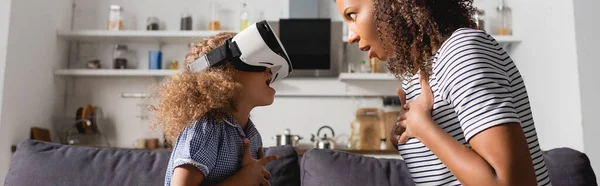 Excited african american babysitter and child in vr headset holding hands on chests while looking at each other, panoramic shot — Stock Photo