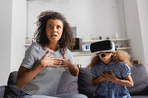 Excited african american nanny and girl in vr headset holding hands on chests while sitting with open mouths — Stock Photo