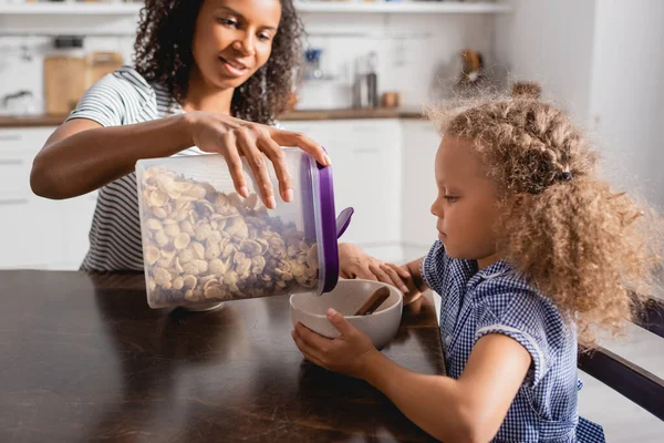 Focalizzazione selettiva della donna afro-americana versando cornflakes dal contenitore nella ciotola vicino alla figlia — Foto stock